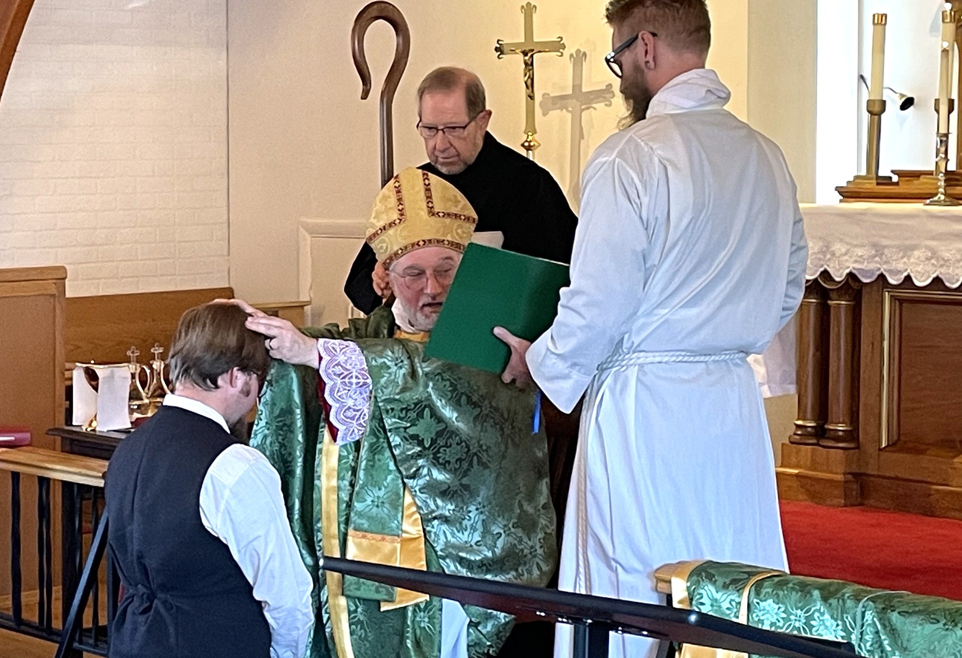 Bishop Fodor, seated in mitre and green vestments, lays his hand on the head of a kneeling candidate while Fr. Nicholas Taylor and a server look on
