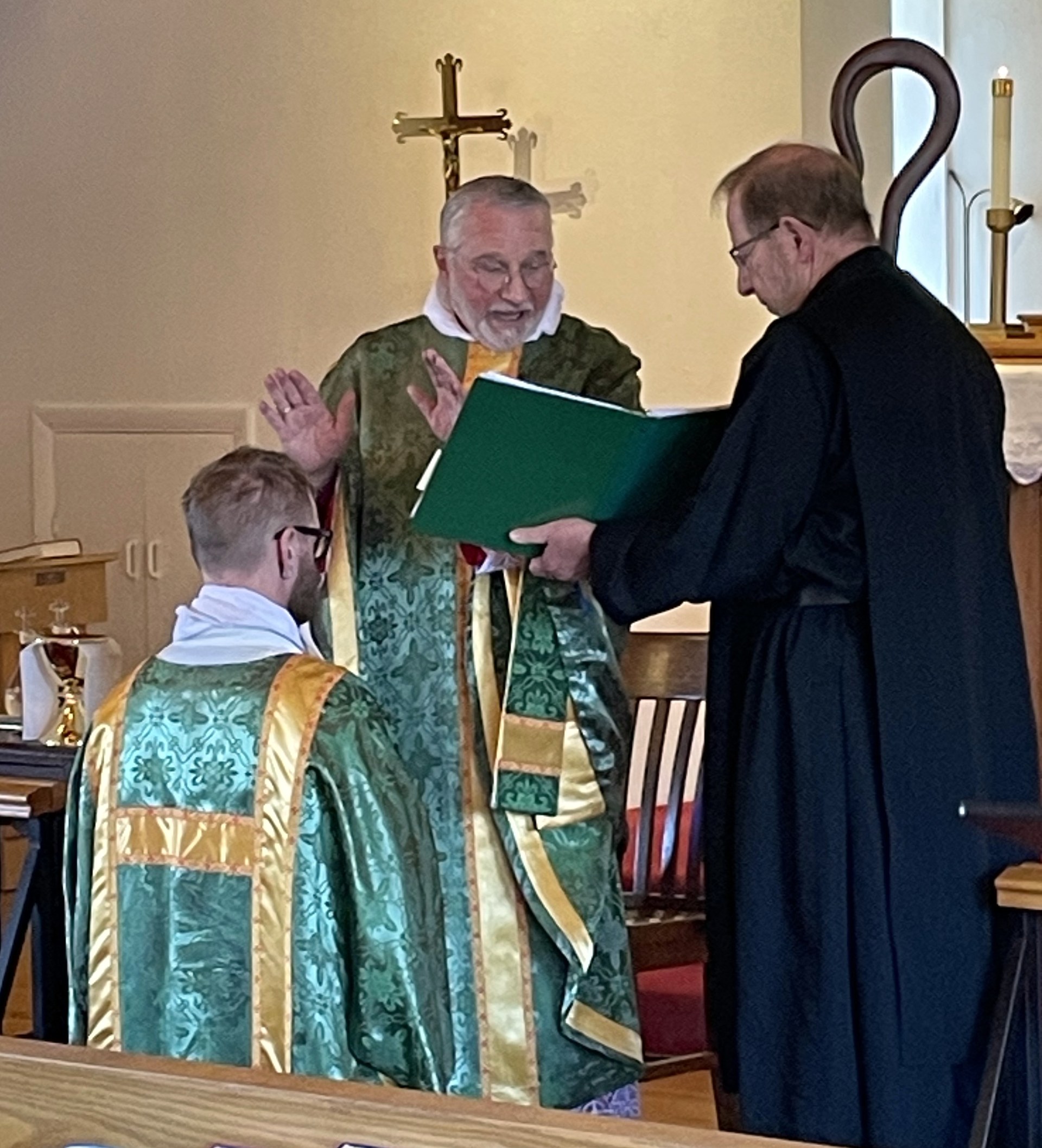 Bishop Fodor, hands raised in prayer, stands over Ryan Steffenhagen kneeling in a green tunicle while Fr. Nicholas Taylor holds the book